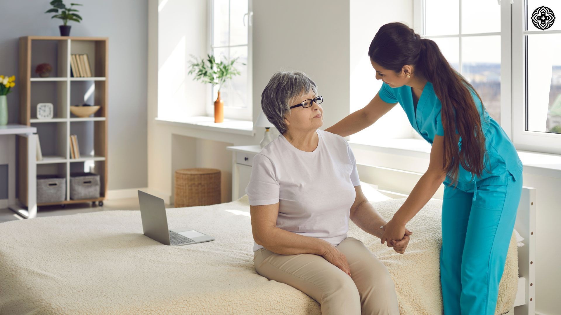 Nurse assisting elderly woman sitting on a bed in a bright room with a laptop nearby.